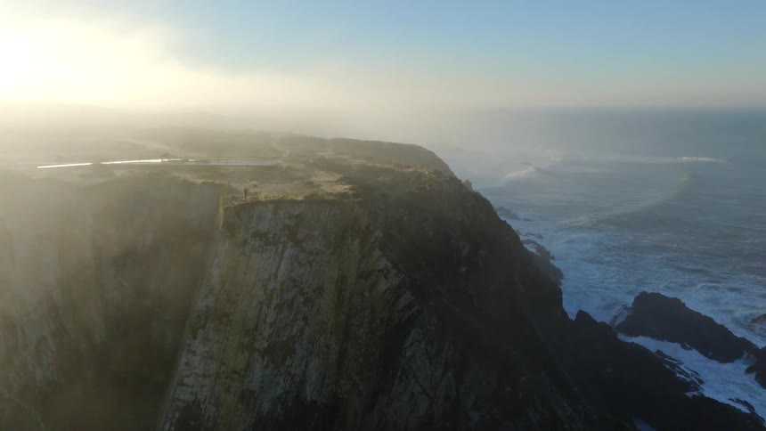An epic aerial view flies through the morning mist to reveal a dramatic, rugged coastline with powerful waves crashing against the cliffs at sunrise