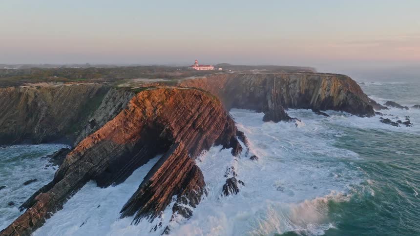 Aerial view pulls back to reveal the stunning and rugged coastline with a lighthouse as powerful ocean waves crash against the dramatic layered cliffs at sunset