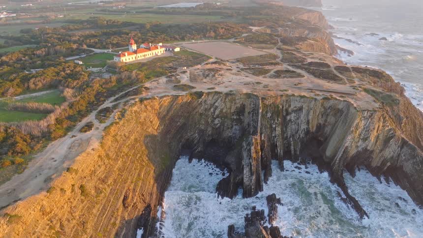 An aerial view pulls back from the stunning Cabo Sardao lighthouse, revealing the rugged cliffs and powerful ocean waves at golden hour