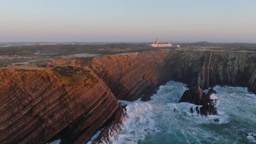 An epic aerial shot reveals the Cabo Sardão lighthouse standing guard over dramatic, layered cliffs as the ocean churns below at sunset