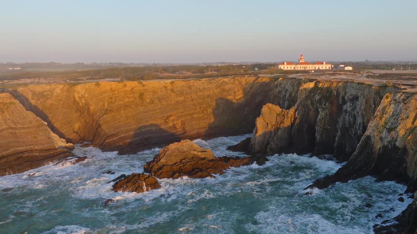 An aerial view pulls back to reveal the stunning Cabo Sardao Lighthouse perched atop dramatic cliffs during a golden sunset