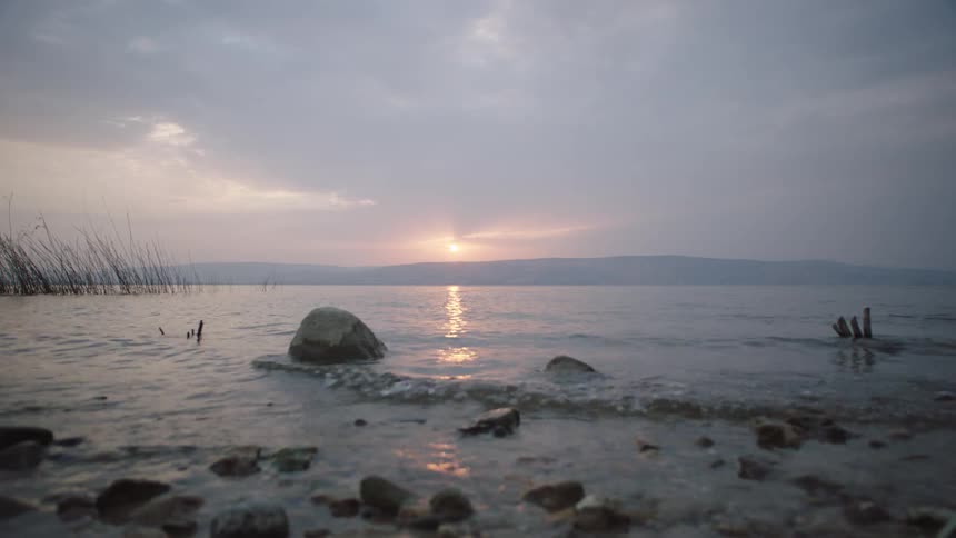 Gentle waves lap the rocky shore of a tranquil lake during a peaceful, cloudy sunrise