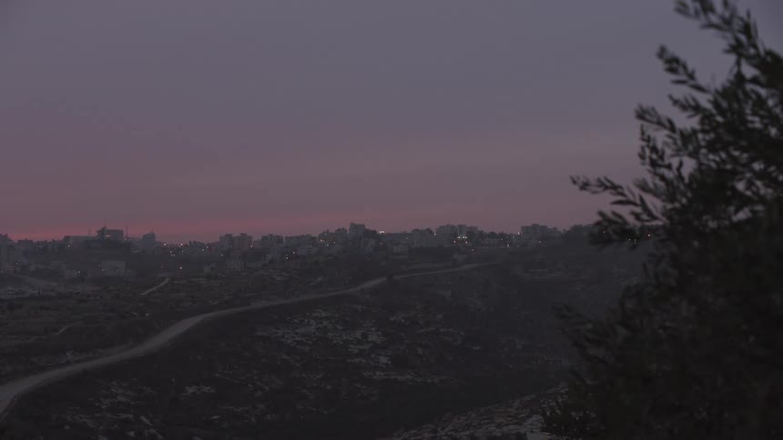 A tranquil view of a distant hillside city with glowing lights under a pink and purple twilight sky