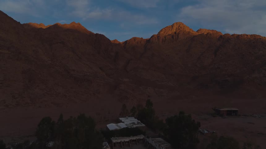 A slow aerial ascent over a quiet desert encampment towards sunlit mountain peaks at sunrise