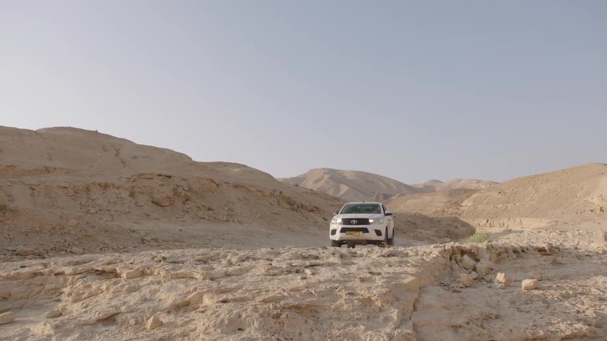 A white 4x4 pickup truck slowly drives over a rocky off-road trail through a vast desert mountain landscape