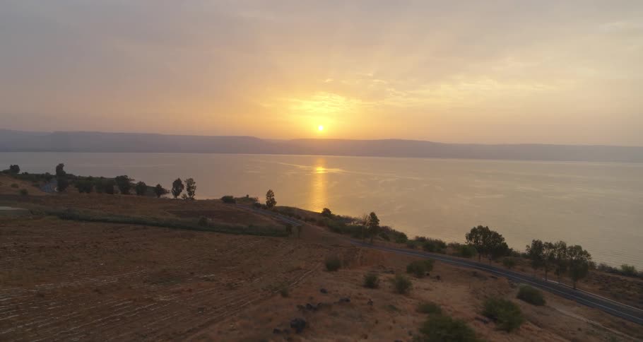 A serene aerial shot captures the golden sunrise reflecting on a vast, calm lake, with a winding road tracing the arid shoreline