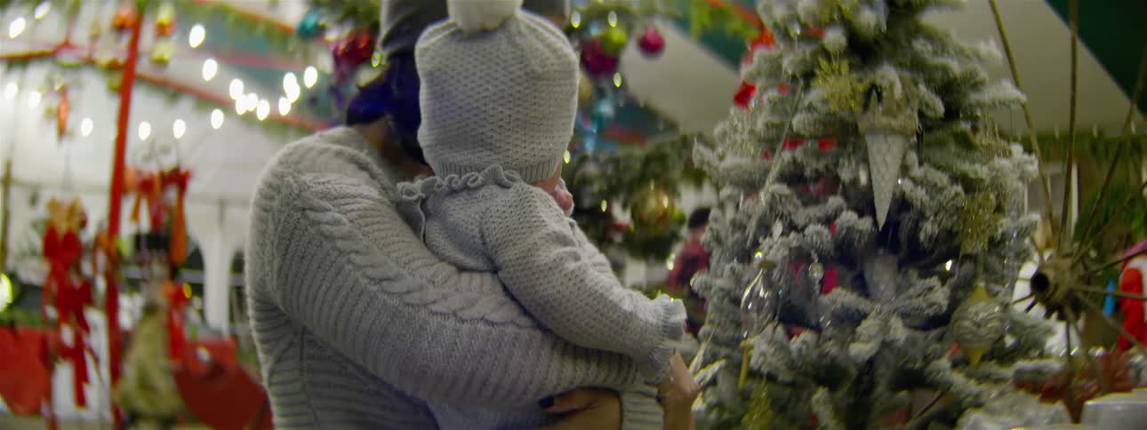 Woman holding baby is looking at decorated christmas tree in store