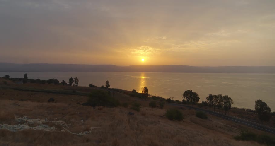 A slow aerial pullback reveals a tranquil sunrise over a vast lake, its golden light reflecting on the calm water and illuminating the dry, grassy hillsides