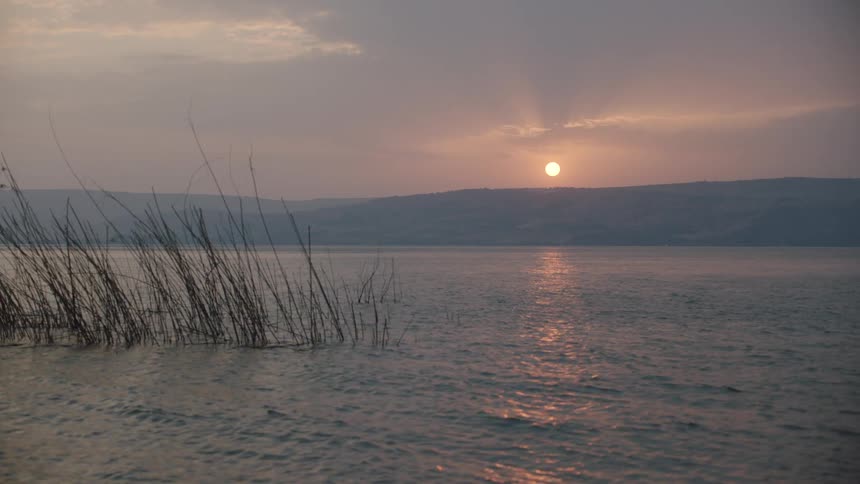 A tranquil sunset over a lake reflects on the calm water with reeds in the foreground and hills in the distance