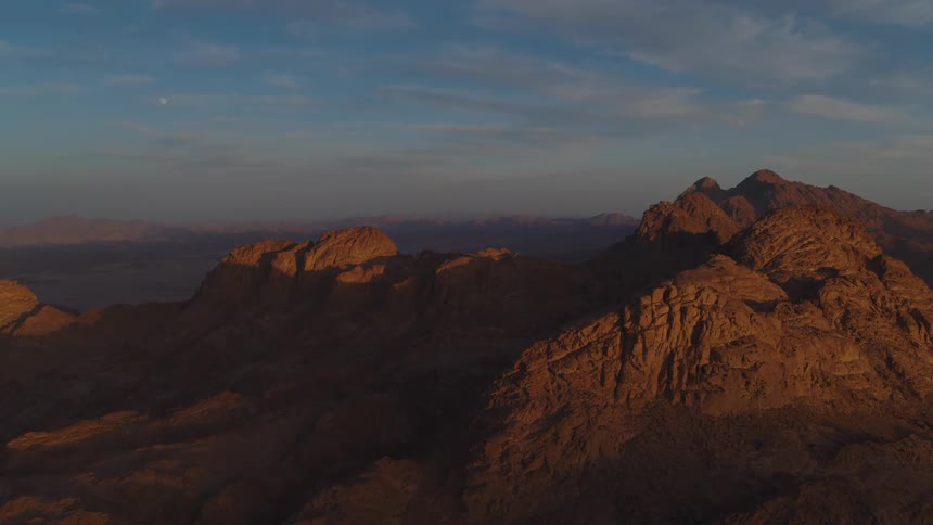 A vast desert mountain range glows in the golden light of sunrise as a full moon sets in the blue sky