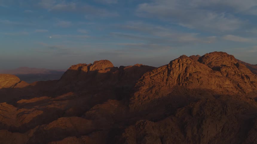 A majestic, rocky desert mountain range glows in the golden light of sunset under a clear blue sky
