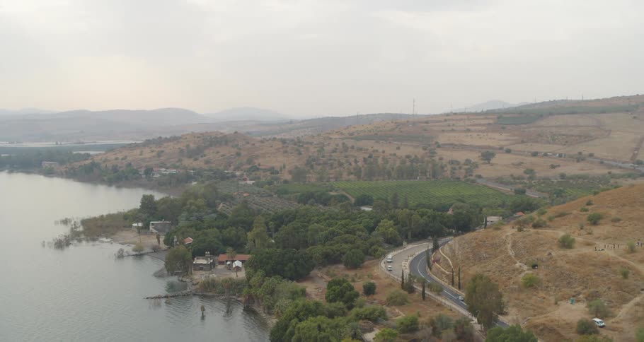 A beautiful aerial shot of the Sea of Galilee coastline, showing a winding road through a landscape of green trees and arid, rolling hills