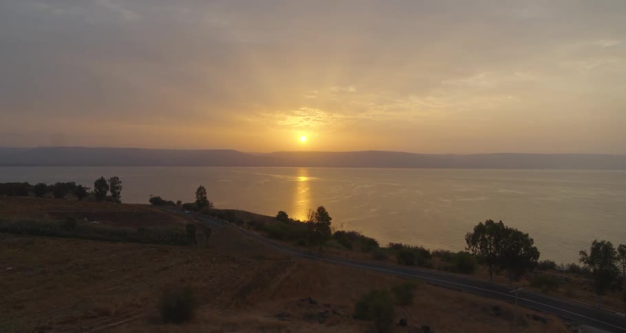 An aerial view shows a car driving along a coastal road next to a vast, calm lake during a beautiful golden sunrise