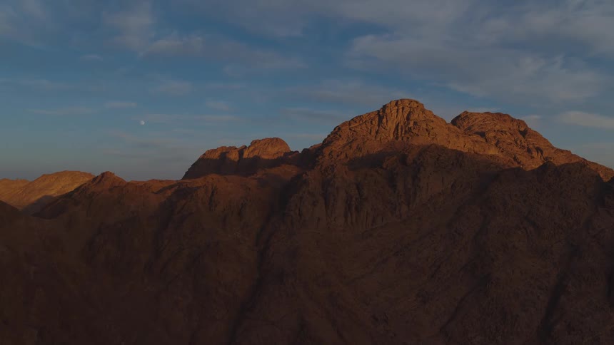 The moon is visible in the sky over a vast, rocky mountain range as the golden light of sunrise begins to illuminate the peaks