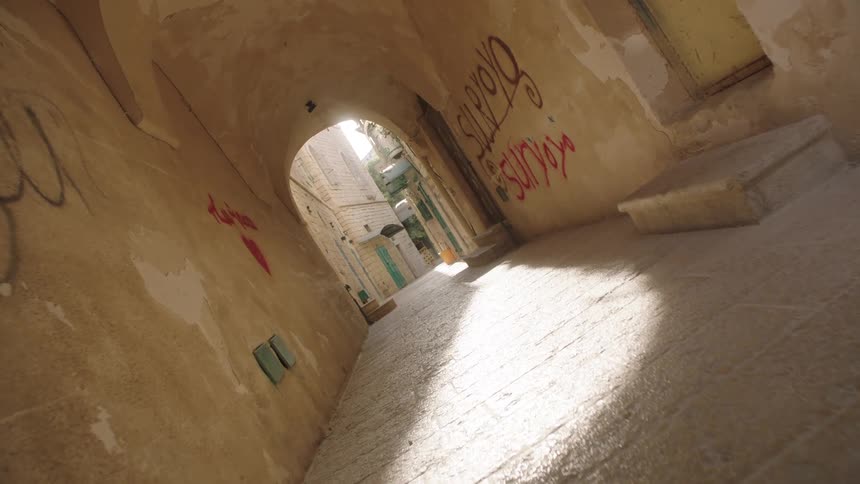 A slow push-in shot down a narrow, sunlit, graffiti-covered stone alleyway in an ancient city
