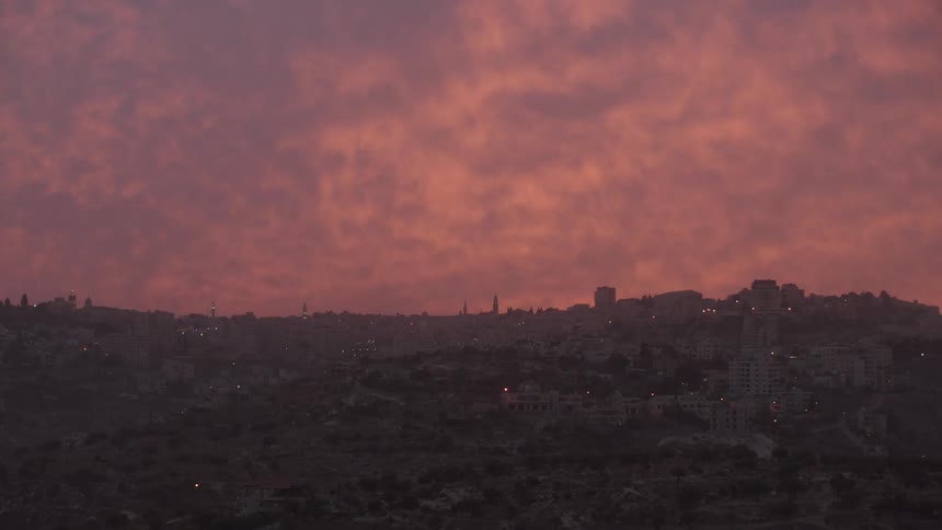 A dramatic pink and orange sunset fills the cloudy sky over a historic city built on rolling hills