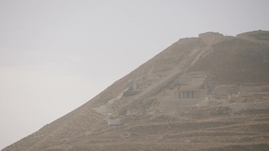 An ancient necropolis or archeological site built into a barren, hazy mountainside slowly comes into focus