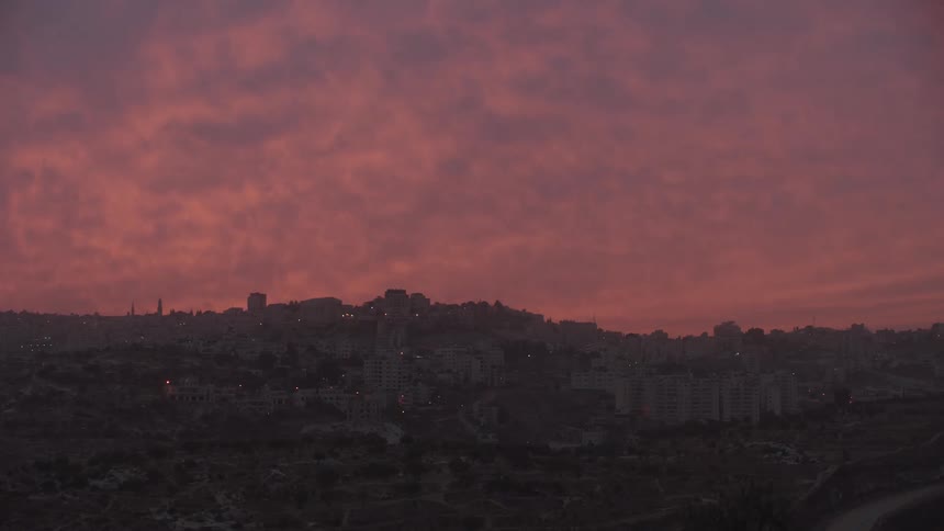 A vibrant pink and orange sunset fills the sky with clouds over the silhouette of a city nestled in the hills