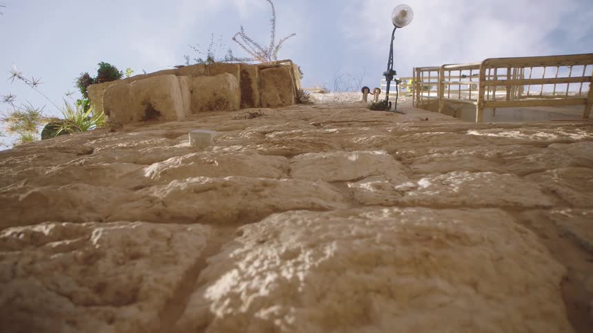 Low-angle tracking shot reveals the texture of a sunlit, ancient stone wall against a bright blue sky