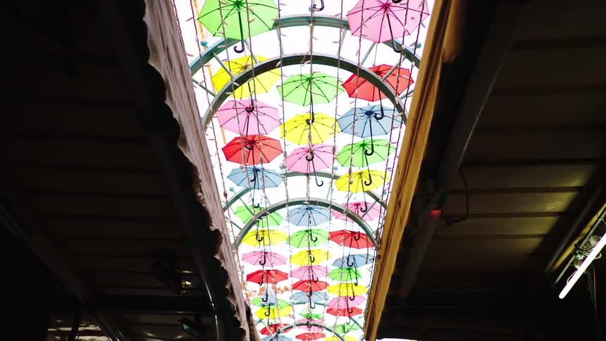 Looking up at a canopy of vibrant, colorful umbrellas suspended over a dark and narrow market street