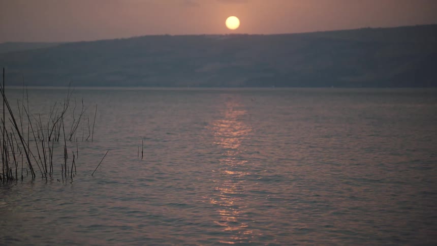 A slow tilt reveals a beautiful sunset reflecting on the calm waters of a vast lake with mountains on the horizon