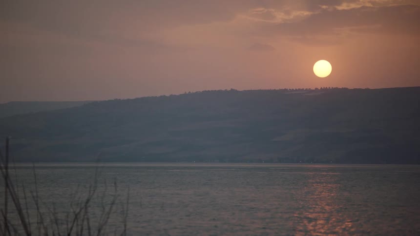 A tranquil sunset casts a warm, orange glow across a vast lake as it slowly dips behind distant, hazy mountains