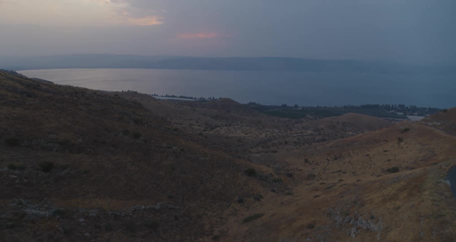 A sweeping view from the arid hills reveals the expansive Sea of Galilee under a dusky, cloudy sky