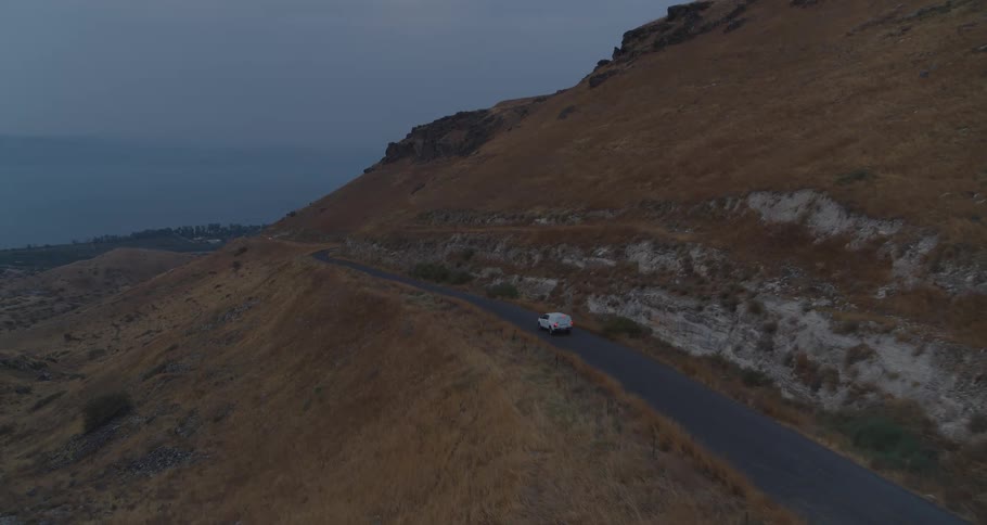 An aerial shot follows a lone white van driving along a winding mountain road next to the sea during a moody twilight evening