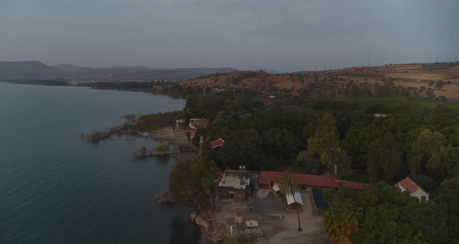 An aerial drone flies over a calm lake towards a verdant shoreline with scattered buildings and distant rolling hills at twilight