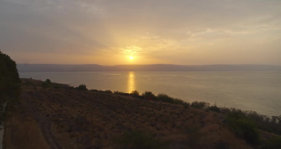 An aerial shot flies forward over the coastline during a beautiful golden sunrise reflecting on a vast, calm lake