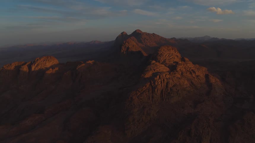 An aerial time-lapse shows the golden morning sun slowly illuminating the peaks of a vast, desolate mountain range