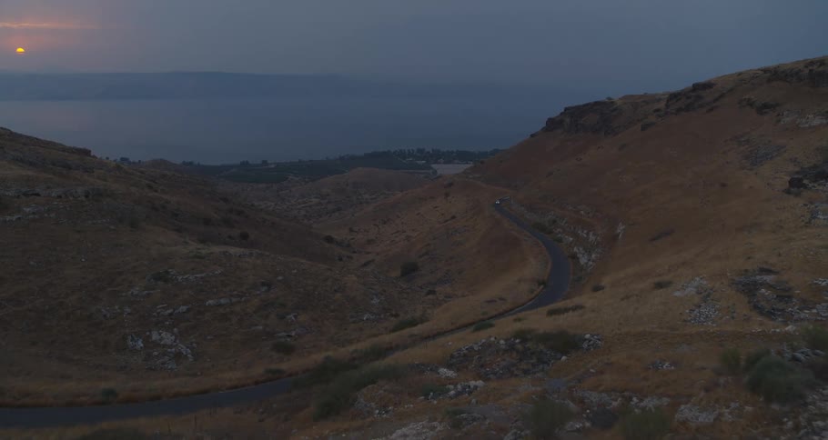An aerial view captures a winding road through a mountain valley overlooking the Sea of Galilee at sunset