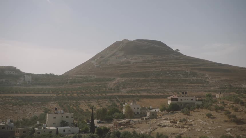 The ancient fortress of Herodium stands prominently over a village in the dry, hilly landscape of the Judean Desert