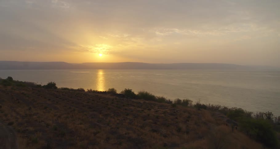 A slow aerial pullback reveals a stunning golden sunrise reflecting over the Sea of Galilee next to a scenic coastal road