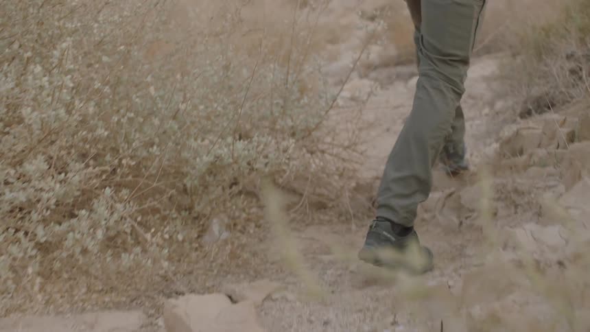 A slow-motion close-up from behind shows a hiker's boots walking along a dusty, rocky desert path