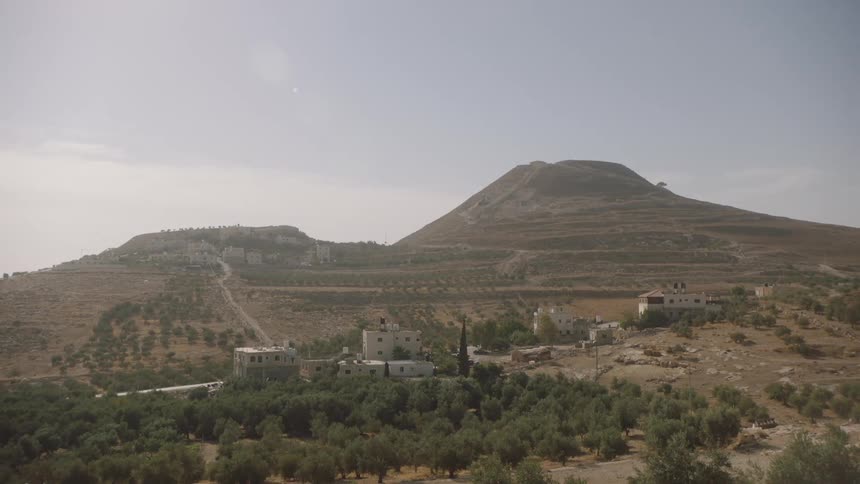 A static wide shot of a village with olive groves nestled among the arid, terraced hills of the Middle East on a hazy day