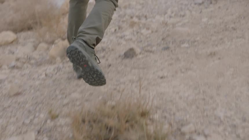 A close-up slow motion shot follows the hiking boots of a person walking up a rocky desert trail