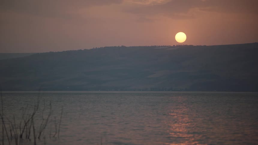 The sun sets over a hazy mountain range, its warm light reflecting on the calm surface of a vast lake