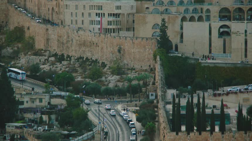 A high-angle view of traffic moving along the ancient walls of the Old City of Jerusalem, tilting up to reveal the historic cityscape