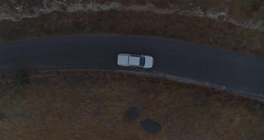 An aerial top-down view follows a white pickup truck driving along a desolate, winding road at dusk