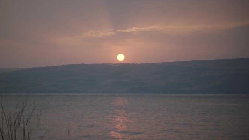 A serene sunset casts a warm glow over a vast lake, with distant mountains silhouetted against the hazy sky