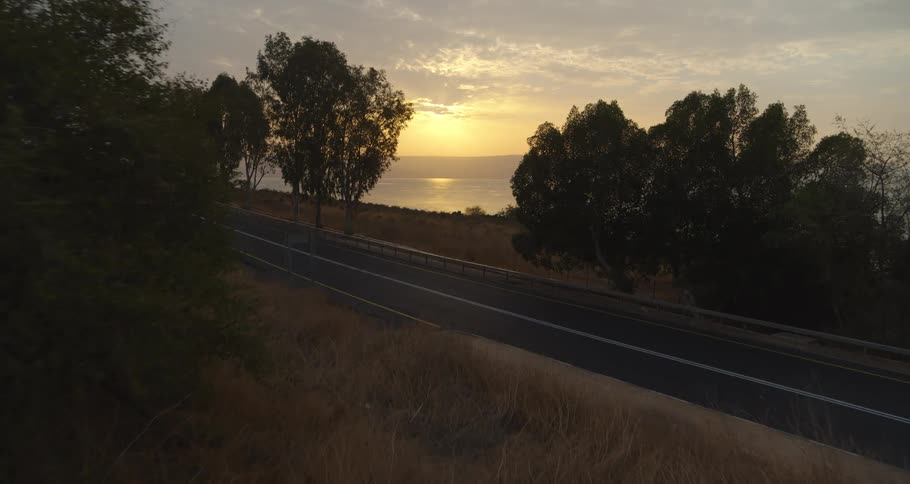 An aerial shot reveals a truck driving on a coastal highway next to a lake during a beautiful sunrise