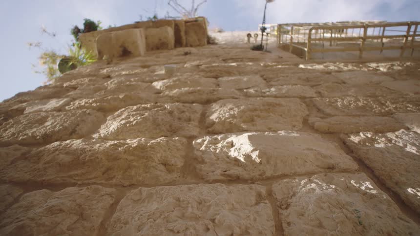 A low-angle view slowly tilts up the weathered, sunlit stones of an ancient wall in a historic city