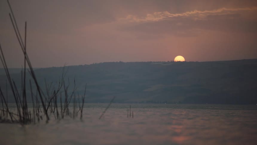 The sun sets behind hazy hills, casting a warm glow over a serene lake with reeds in the foreground