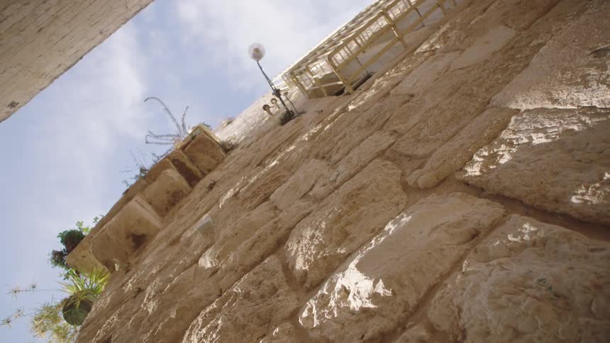 A slow, low-angle dolly shot follows the sun-drenched texture of an ancient stone wall in an old city alley