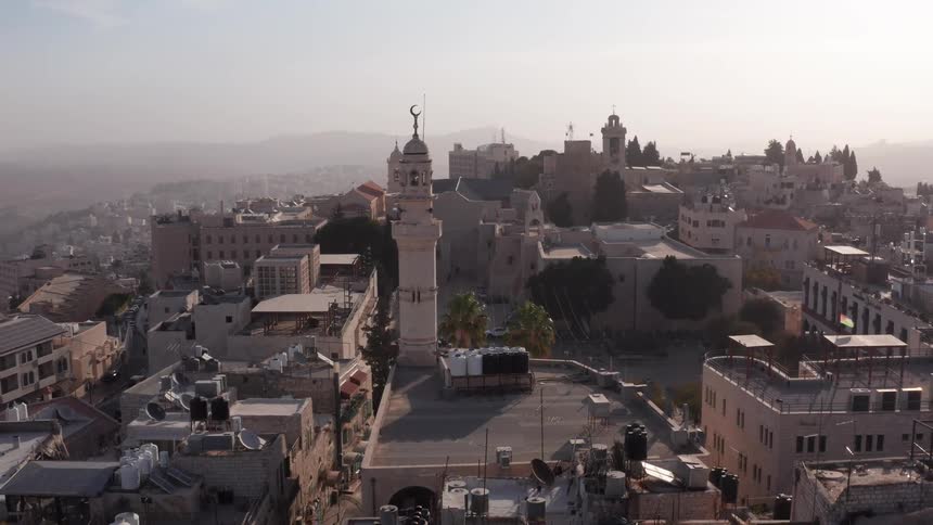 A slow aerial pullback reveals the historic city of Bethlehem and a central mosque minaret during a hazy sunrise