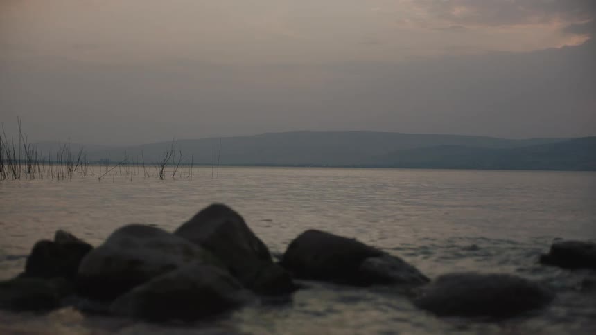 Gentle waves ripple on a tranquil lake at twilight with rocks on the shoreline and hazy mountains in the background