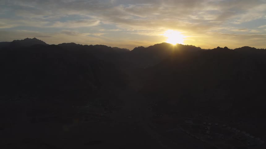 A serene aerial view shows the sun rising behind a silhouetted mountain range over a desert settlement