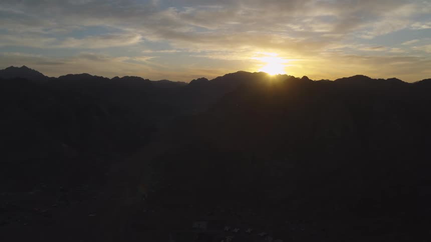 A static aerial view shows the sun breaking over the peaks of a vast, silhouetted mountain range at dawn