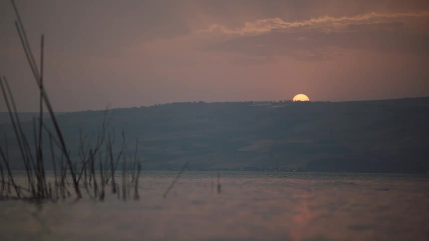 The setting sun casts a gentle glow over a calm lake as it disappears behind a distant mountain, with reeds in the foreground
